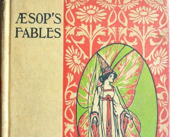 May include: Vintage hardcover book titled "Aesop's Fables." The cover features a cream-colored background with the title in black. A red and cream floral design borders a green oval with an illustration of a woman with butterfly wings.
