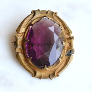 May include: An oval brooch featuring a large, faceted deep purple stone set within a textured, antique-toned brass frame. The metal setting has a decorative, scalloped edge with prongs securing the central gemstone in place.