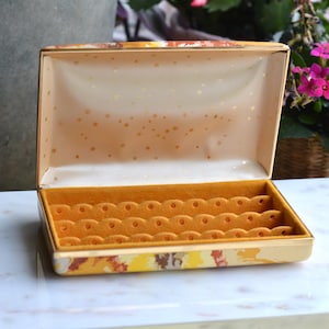May include: Open vintage jewelry box with a patterned exterior in shades of yellow, orange, and brown. The interior is lined with a soft, golden-yellow material with slots. The box is open, revealing the interior, and sits on a white surface.