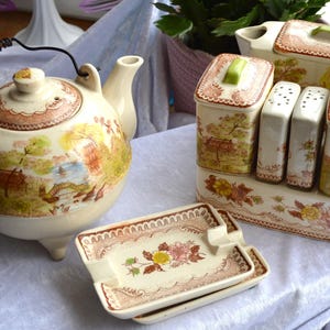May include: A vintage cream-colored tea set with a landscape design. The set includes a teapot, a sugar bowl, two salt and pepper shakers, two canisters, and two ashtrays. The set has a brown and pink floral border and green accents.