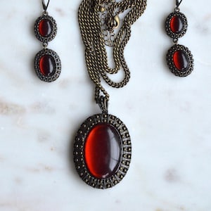 May include: A vintage jewelry set featuring a necklace and matching earrings. The set includes a bronze-toned chain and pendant with a large oval, red gemstone. The earrings have a similar design with smaller red stones, all set in a detailed, ornate frame.