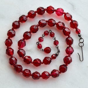 May include: A necklace composed of faceted, translucent deep red beads arranged in a spiral pattern. The beads are strung on a thin wire, and the piece features a dark metal hook-and-eye clasp mechanism at one end to secure the necklace.