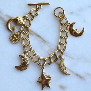 May include: A gold-toned charm bracelet featuring a toggle clasp and multiple celestial-themed charms, including various crescent moons, a sun with a face, a textured star, and a small round globe charm, all attached to a double-link chain.