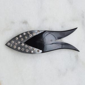 May include: A black hair accessory shaped like a stylized fish, featuring a smooth, glossy finish on the tail section and a curved front section adorned with multiple clear, round rhinestones embedded in a dark, textured surface.