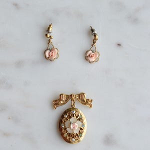 Puede incluir: Un conjunto de joyas vintage con un colgante de medall&oacute;n dorado con una rosa rosa y detalles de pedrer&iacute;a, y pendientes a juego con detalles de rosas rosas. El conjunto est&aacute; sobre una superficie de m&aacute;rmol blanco.