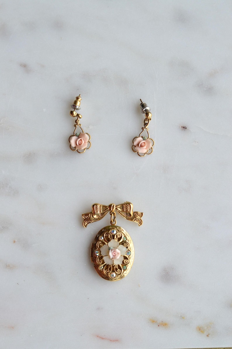 Puede incluir: Un conjunto de joyas vintage con un colgante de medall&oacute;n dorado con una rosa rosa y detalles de pedrer&iacute;a, y pendientes a juego con detalles de rosas rosas. El conjunto est&aacute; sobre una superficie de m&aacute;rmol blanco.