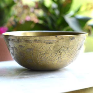 May include: A round brass bowl featuring an etched decorative pattern around the exterior rim and body. The metal surface displays a weathered, golden-brown patina with visible dark markings and intricate line work engraved into the metal.
