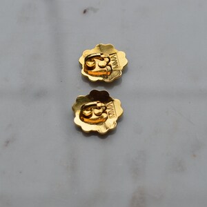 May include: Two gold-colored flower-shaped earrings with a scalloped edge. The earrings have a post back and the letters "MMA" are visible on the back of each earring. The earrings are on a light gray surface.