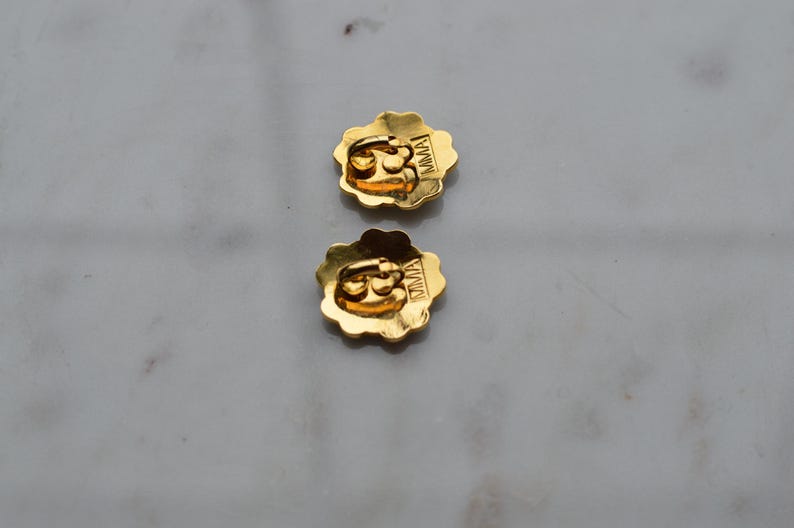 May include: Two gold-colored flower-shaped earrings with a scalloped edge. The earrings have a post back and the letters "MMA" are visible on the back of each earring. The earrings are on a light gray surface.