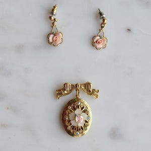 Puede incluir: Un conjunto de joyas vintage con un broche y pendientes a juego en tono dorado. El broche es un relicario ovalado con un lazo, una rosa rosa central y cristales transparentes y azules. Los pendientes tienen colgantes de rosas rosas.