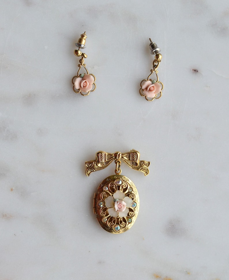 Puede incluir: Un conjunto de joyas vintage con un broche y pendientes a juego en tono dorado. El broche es un relicario ovalado con un lazo, una rosa rosa central y cristales transparentes y azules. Los pendientes tienen colgantes de rosas rosas.