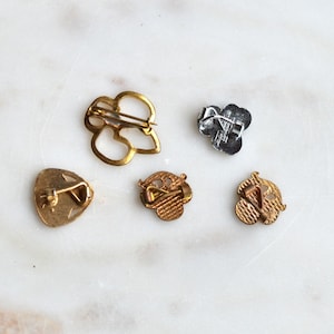 May include: Five metal pins shown from the back, including one large gold-tone open-work pin, one small silver-tone pin, and three small gold-tone pins with textured surfaces. Each pin features a metal pin-back mechanism for attachment.