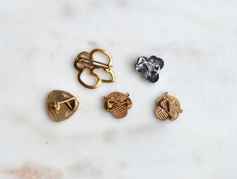 May include: Five metal pins shown from the back, including one large gold-tone open-work pin, one small silver-tone pin, and three small gold-tone pins with textured surfaces. Each pin features a metal pin-back mechanism for attachment.