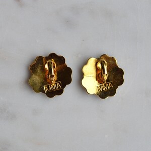 May include: Two gold-toned clip-on earrings with a scalloped edge design. The backs of the earrings have a clip mechanism and the letters "MMA" stamped on them. The earrings are on a marble surface.