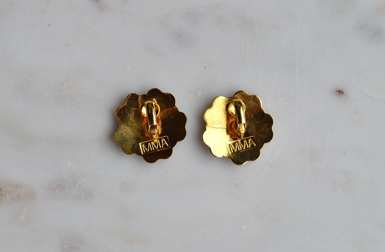 May include: Two gold-toned clip-on earrings with a scalloped edge design. The backs of the earrings have a clip mechanism and the letters "MMA" stamped on them. The earrings are on a marble surface.