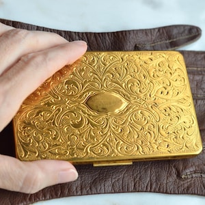 May include: A rectangular gold-colored metal case featuring an intricate embossed floral and scrollwork pattern across the surface, with a smooth oval-shaped center panel, resting on top of a dark brown leather glove.