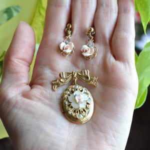 Puede incluir: Un conjunto de joyas vintage con un broche dorado y pendientes a juego. El broche es de forma ovalada con un dise&ntilde;o floral y un detalle de lazo. Los pendientes y el broche tienen detalles florales rosas y blancos.