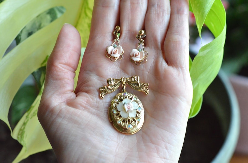 Puede incluir: Un conjunto de joyas vintage con un broche dorado y pendientes a juego. El broche es de forma ovalada con un dise&ntilde;o floral y un detalle de lazo. Los pendientes y el broche tienen detalles florales rosas y blancos.
