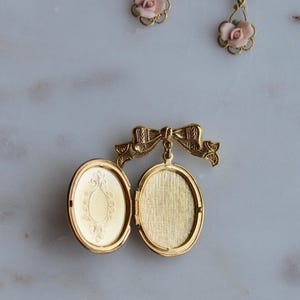 Puede incluir: Medall&oacute;n ovalado dorado abierto, con un dise&ntilde;o ornamentado en el interior. Est&aacute; sujeto a un cierre en forma de lazo. Dos pares de pendientes de rosas rosas se ven en el fondo.