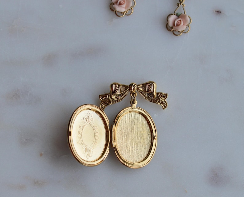 Puede incluir: Medall&oacute;n ovalado dorado abierto, con un dise&ntilde;o ornamentado en el interior. Est&aacute; sujeto a un cierre en forma de lazo. Dos pares de pendientes de rosas rosas se ven en el fondo.