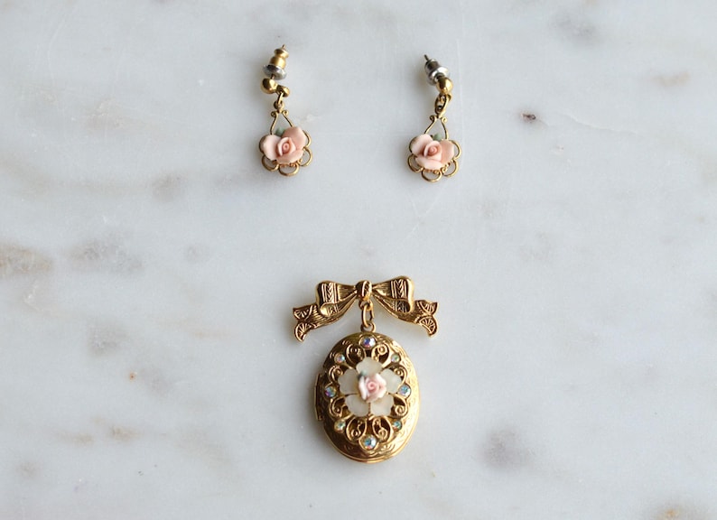 May include: A set of vintage jewelry, including a gold-toned locket pendant with a pink rose and clear crystal accents, and a pair of matching earrings. The earrings and pendant feature a floral design.
