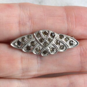 May include: A silver-toned metal brooch featuring a symmetrical, fan-like shape with a series of small, dark, faceted stones embedded throughout the surface. The design includes intricate engraved lines and a pointed, elongated wing-like structure.