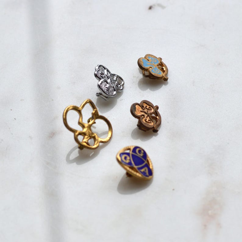 May include: A collection of five small metal pins featuring various designs, including a silver-toned clover, a light blue enamel clover, a plain bronze-toned clover, a gold-toned open-work clover, and a triangular gold-toned pin with blue enamel and the letters GCA.