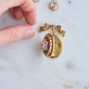 Puede incluir: Medall&oacute;n dorado con dise&ntilde;o floral y una peque&ntilde;a rosa rosa. El medall&oacute;n est&aacute; abierto, revelando un interior dorado. Tambi&eacute;n se ven un detalle de lazo a juego y dos pendientes con detalles de rosas.