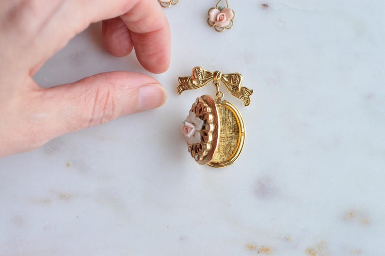 Puede incluir: Medall&oacute;n dorado con dise&ntilde;o floral y una peque&ntilde;a rosa rosa. El medall&oacute;n est&aacute; abierto, revelando un interior dorado. Tambi&eacute;n se ven un detalle de lazo a juego y dos pendientes con detalles de rosas.