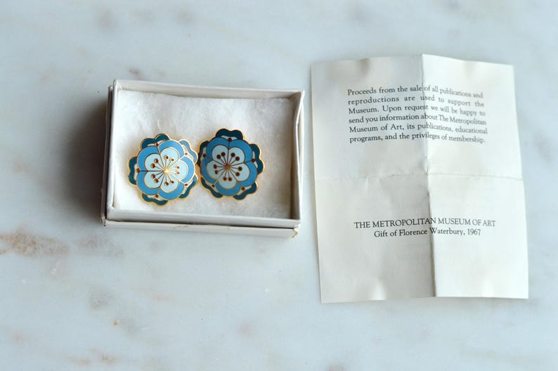 May include: A pair of floral-shaped earrings in a white box. The earrings feature a light blue and white design with gold trim. A paper with text from The Metropolitan Museum of Art is next to the box.