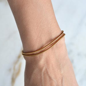 May include: A gold-toned snake chain bracelet featuring a flexible, rounded design with a smooth, continuous texture. The bracelet wraps around the wrist in two parallel loops, showcasing a metallic finish and a slender, uniform link construction.