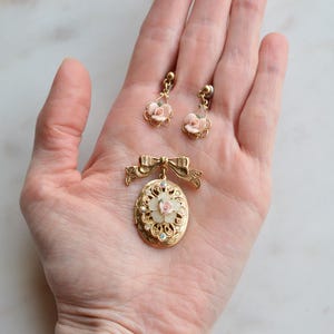 Puede incluir: Un conjunto de joyas vintage con un colgante dorado con lazo y medall&oacute;n ovalado, y pendientes a juego. Los pendientes y el colgante tienen delicados detalles de rosas rosas. El conjunto se exhibe sobre una superficie de m&aacute;rmol.