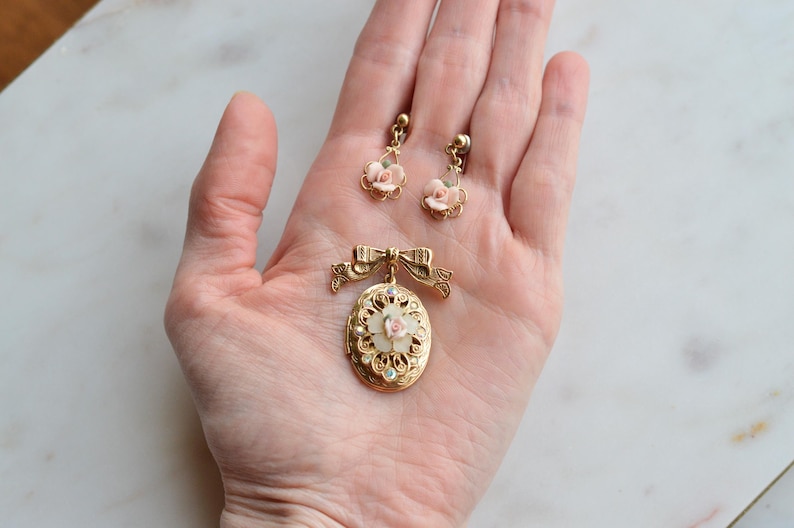 Puede incluir: Un conjunto de joyas vintage con un colgante dorado con lazo y medall&oacute;n ovalado, y pendientes a juego. Los pendientes y el colgante tienen delicados detalles de rosas rosas. El conjunto se exhibe sobre una superficie de m&aacute;rmol.