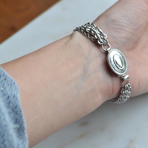 May include: A silver-toned multi-strand chain bracelet featuring a central oval-shaped medallion with a smooth, raised center and a textured, rope-like border. The bracelet consists of several thin, delicate chains connected to the central piece.