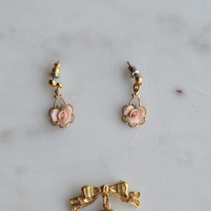 Puede incluir: Un par de pendientes vintage dorados con detalles de rosas rosas. Cada pendiente presenta una peque&ntilde;a rosa dentro de un marco festoneado, suspendida de un poste decorativo. Los pendientes se presentan sobre una superficie de m&aacute;rmol.
