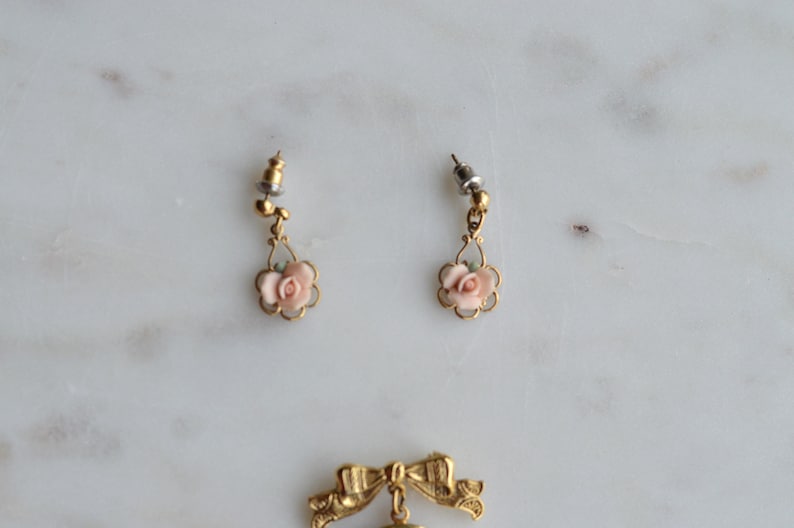Puede incluir: Un par de pendientes vintage dorados con detalles de rosas rosas. Cada pendiente presenta una peque&ntilde;a rosa dentro de un marco festoneado, suspendida de un poste decorativo. Los pendientes se presentan sobre una superficie de m&aacute;rmol.