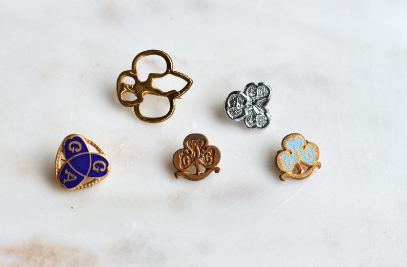 May include: A collection of five metal pins featuring various designs, including a gold-toned open clover outline, a silver-toned textured clover, a blue triangular pin with the letters G A, and two small gold-toned clover pins with blue accents.