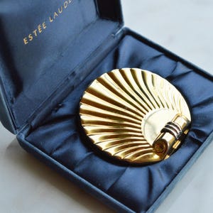 May include: A vintage gold-toned compact mirror with a fan-like design, resting in a navy blue velvet case. The case is open, revealing the compact. The brand name "ESTEE LAUDE" is printed in gold lettering on the case.