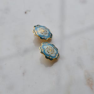 May include: Two floral-shaped earrings with a light blue and white enamel design and gold-colored trim. The earrings are set against a white marble surface. The earrings have a delicate, feminine aesthetic.