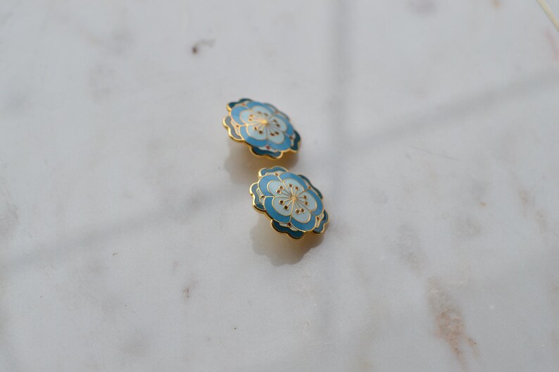 May include: Two floral-shaped earrings with a light blue and white enamel design and gold-colored trim. The earrings are set against a white marble surface. The earrings have a delicate, feminine aesthetic.