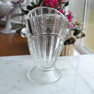 May include: A clear, vintage glass vase with a textured, ribbed design. The vase has a flared top with two small handles and a decorative base. It is sitting on a white surface, with flowers in the background.