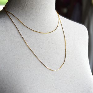 May include: A delicate gold-toned chain necklace featuring a double-layered design. The necklace consists of two thin, smooth metallic strands that drape around the neck, creating a layered appearance with a polished, reflective finish throughout.
