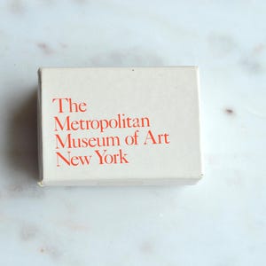 May include: A rectangular, off-white box with the text "The Metropolitan Museum of Art New York" printed in red. The box is centered on a white and gray marble surface.