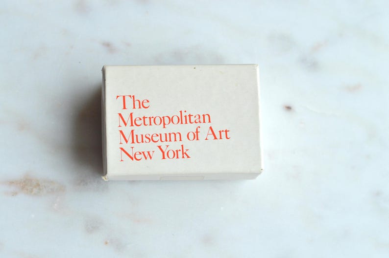 May include: A rectangular, off-white box with the text "The Metropolitan Museum of Art New York" printed in red. The box is centered on a white and gray marble surface.
