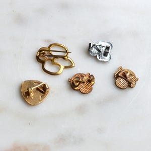 May include: Five metal pins shown from the back, including one large gold-toned openwork pin, one silver-toned pin, and three smaller gold-toned pins with textured backs and pin-back closures, displaying various shapes and metallic finishes.