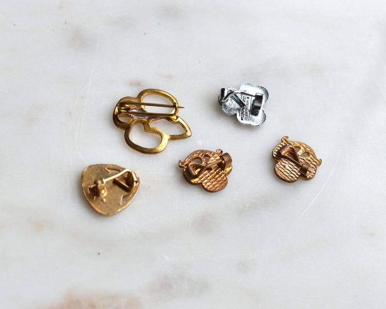 May include: Five metal pins shown from the back, including one large gold-toned openwork pin, one silver-toned pin, and three smaller gold-toned pins with textured backs and pin-back closures, displaying various shapes and metallic finishes.