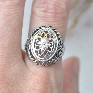 May include: A silver-toned ring featuring an oval-shaped face with an intricate openwork filigree design. The center of the oval displays a detailed floral motif, while the band and the surrounding frame are crafted with ornate, swirling metal patterns.
