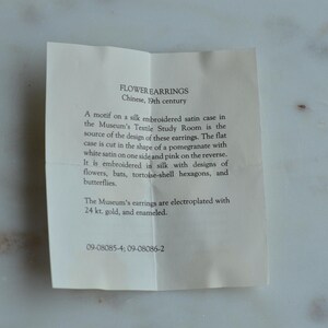 May include: A folded paper with text describing "FLOWER EARRINGS" from 19th century China. The text details the design source, a silk embroidered satin case, and the materials used, including gold and enamel.