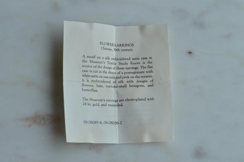 May include: A folded paper with text describing "FLOWER EARRINGS" from 19th century China. The text details the design source, a silk embroidered satin case, and the materials used, including gold and enamel.