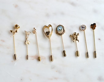 May include: A collection of eight gold-tone stick pins featuring various decorative tops including a rose, a bow, a heart, a blue floral cameo with pearls, a dancing figure, a circular floral motif, and a flower shape, all arranged in a row.